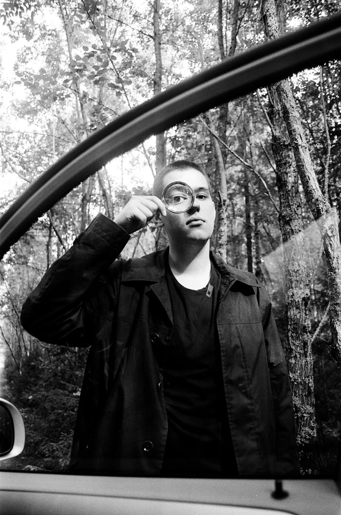 Black and white photo of a man holding a magnifier in a tranquil forest, seen through a car window.
